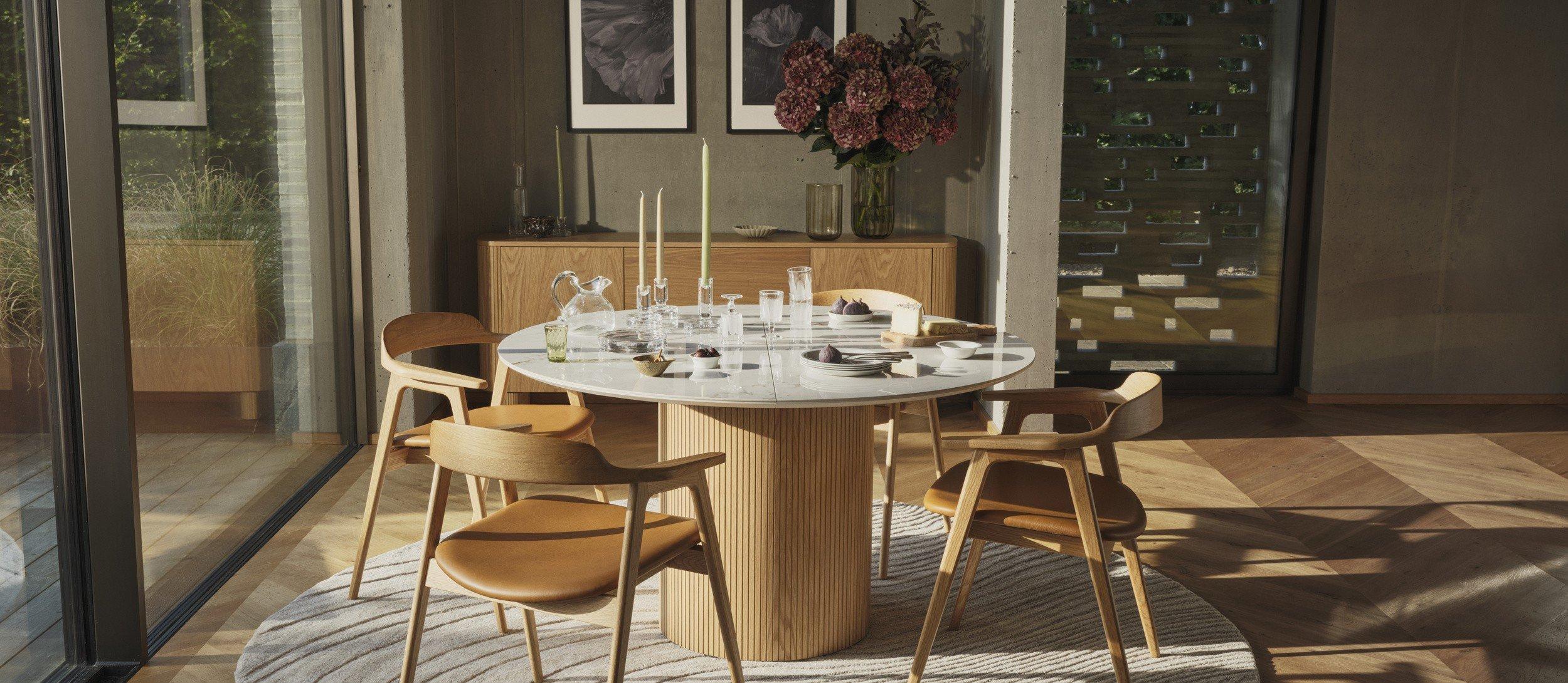 Moderná jedáleň s okrúhlym dreveným stolom Santiago, štyrmi koženými stoličkami Seoul, textúrovaným kobercom a príborníkom s obrazom v ráme s kvetmi.